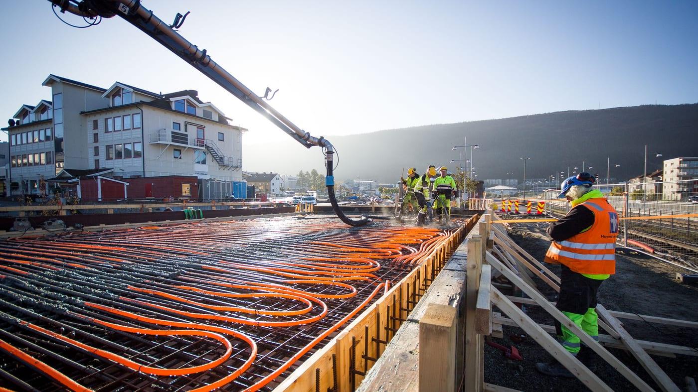 A concrete pump pouring a slab over orange underfloor heating pipes while a worker in hi-vis guides it