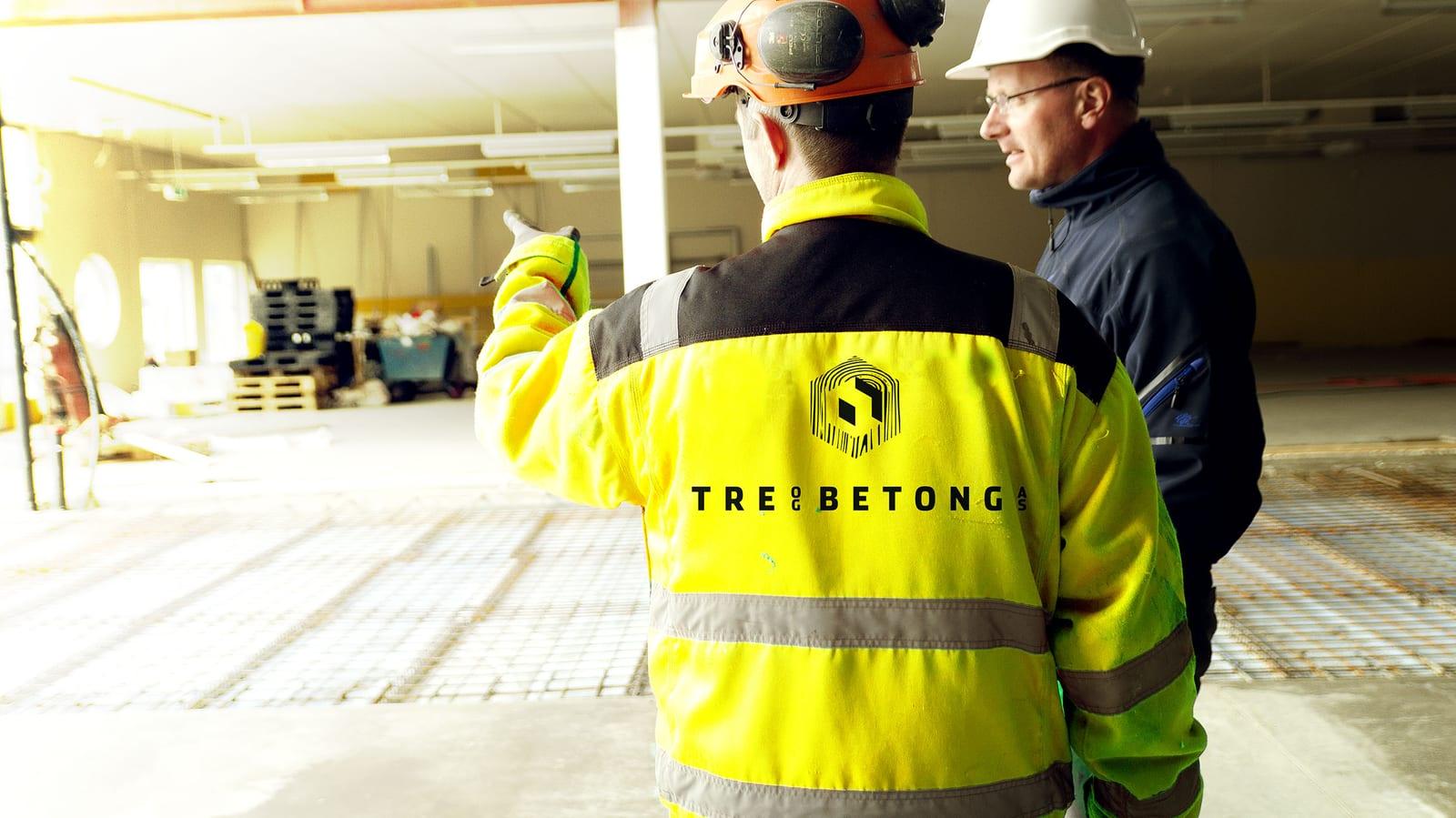 Two men in Tre og Betong hi-vis jackets and hard hats inspecting a large empty floor inside a building