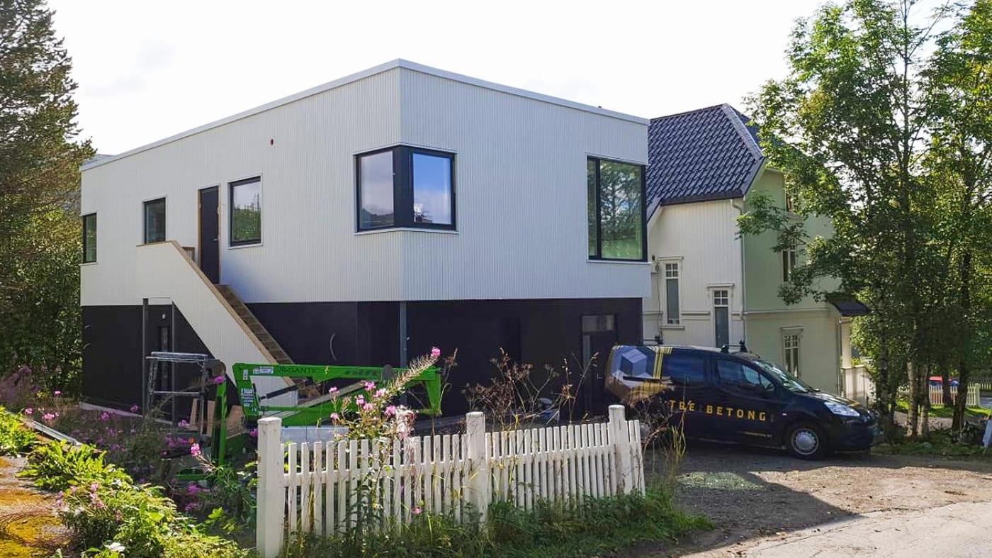 A white-clad extension with a garage below and a flat above, beside an older timber house, with a Tre og Betong van parked in front