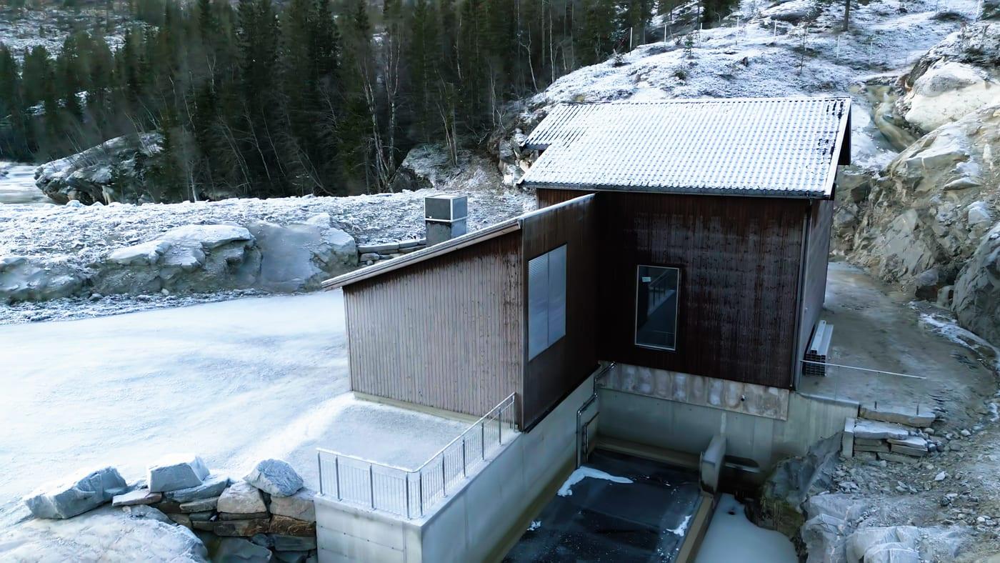 A small timber-clad power station building by a frozen river, with a concrete intake