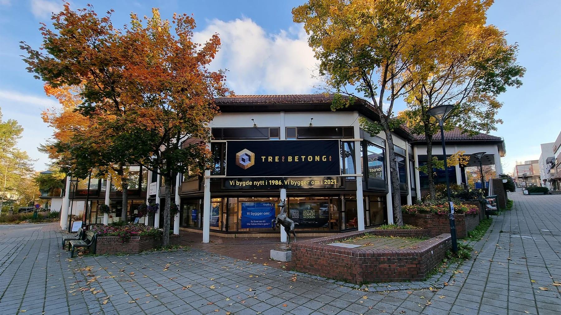 A Tre og Betong banner across the front of the Sparebank 1 Helgeland building, with autumn trees in the square